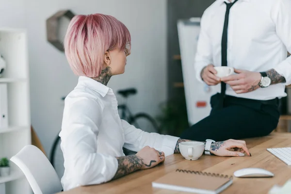 Junge Geschäftsfrau mit Tätowierungen schaut männlichen Kollegen an, der mit einer Tasse Kaffee am Tisch sitzt — Stockfoto