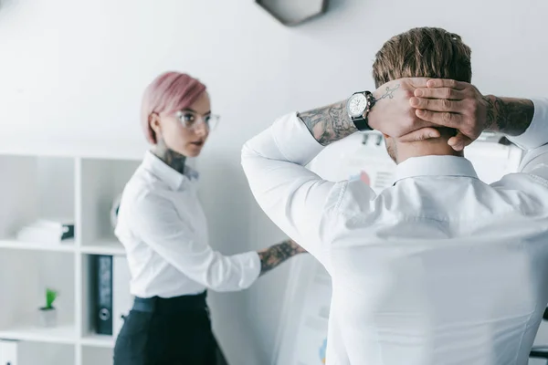 Vista trasera del hombre de negocios de pie con las manos detrás de la cabeza y mirando a una colega apuntando a la pizarra blanca - foto de stock