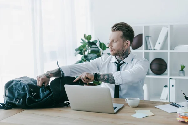 Young tattooed businessman opening backpack while working with laptop at workplace — Stock Photo