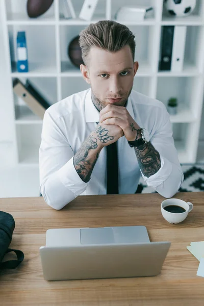 Young tattooed businessman looking at camera while working with laptop at workplace — Stock Photo