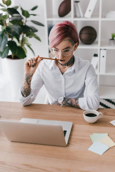 Joven empresaria con tatuajes sosteniendo lápiz y usando laptop en el lugar de trabajo - foto de stock