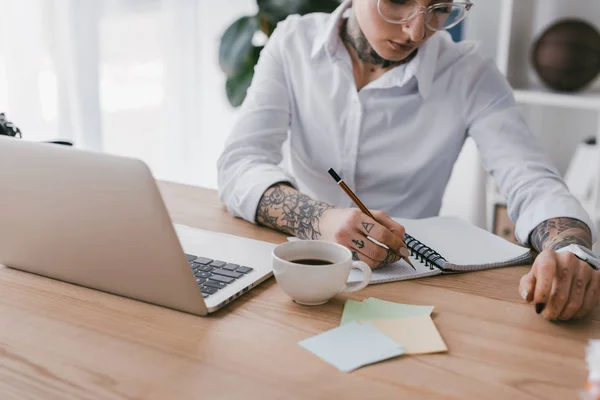 Schnappschuss von junger Geschäftsfrau, die Notizen macht, während sie Laptop am Arbeitsplatz benutzt — Stockfoto