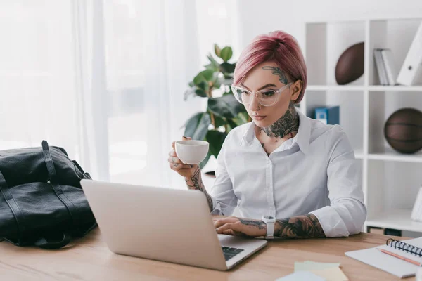 Joven mujer de negocios tatuada sosteniendo taza de café y el uso de la computadora portátil en la oficina - foto de stock