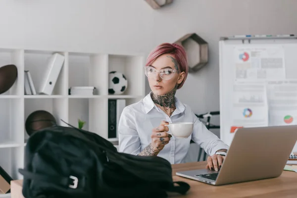 Junge Geschäftsfrau hält eine Tasse Kaffee in der Hand und schaut weg, während sie am Arbeitsplatz Laptop benutzt — Stockfoto