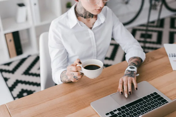 Colpo ritagliato di giovane donna d'affari con tatuaggi in possesso di una tazza di caffè e utilizzando il computer portatile — Foto stock