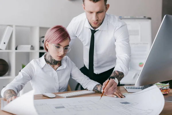 Young tattooed colleagues in formal wear working with blueprint in office — Stock Photo