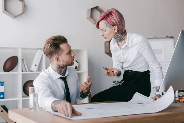 Young tattooed business people talking and looking at each other while working with blueprint in office — Stock Photo