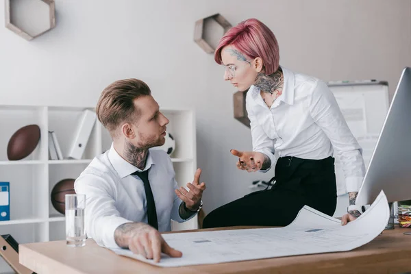 Jóvenes hombres de negocios tatuados hablando y mirándose mientras trabajan con planos en la oficina - foto de stock