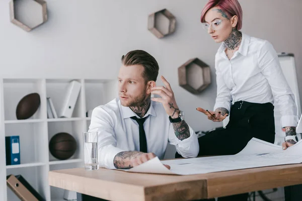 Junge tätowierte Geschäftsleute unterhalten sich während der Arbeit mit Blaupausen im Büro — Stockfoto