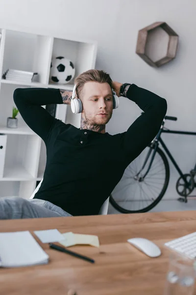 Beau jeune homme tatoué dans des écouteurs assis avec les mains derrière la tête sur le lieu de travail — Photo de stock