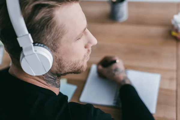 Vista de cerca de un joven empresario con auriculares mirando hacia otro lado mientras toma notas en el lugar de trabajo - foto de stock