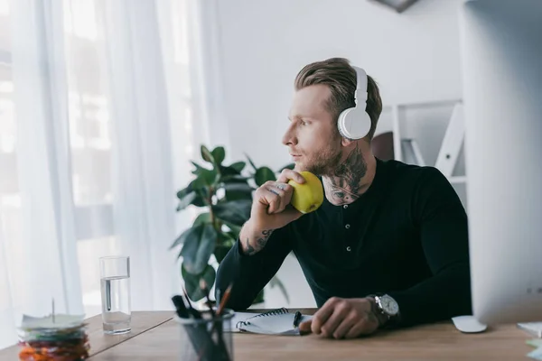 Pensive young businessman in headphones holding apple and looking away at workplace — Stock Photo