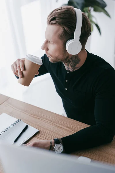 High angle view of young tattooed businessman in headphones drinking coffee from paper cup — Stock Photo