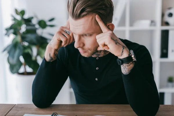 Serious young businessman sitting at table and looking away in office — Stock Photo
