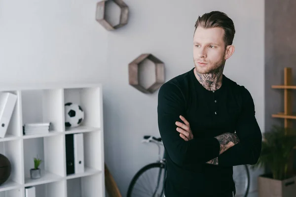 Handsome young tattooed businessman standing with crossed arms and looking away in office — Stock Photo