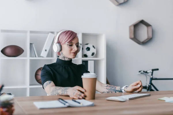 Sonriente joven empresaria escuchando música en auriculares y sosteniendo una taza de papel en el lugar de trabajo - foto de stock