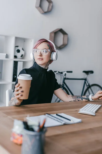 Jovem empresária em fones de ouvido segurando café para ir e olhando para longe no local de trabalho — Fotografia de Stock