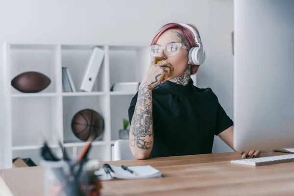 Young tattooed businesswoman in headphones looking away at workplace — Stock Photo