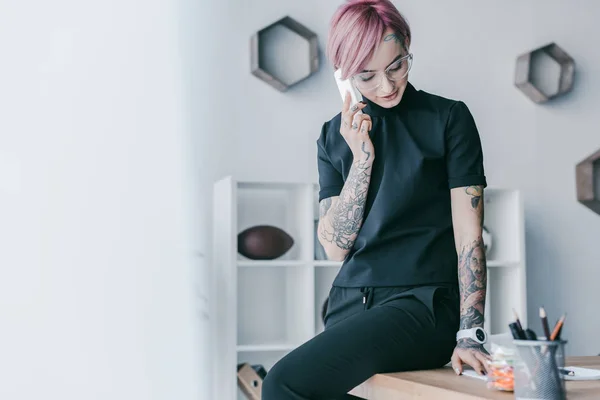 Jeune femme d'affaires souriante avec des tatouages parlant par smartphone sur le lieu de travail — Photo de stock