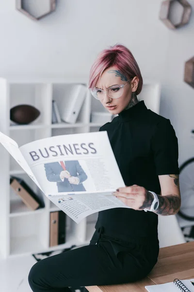 Seria joven mujer de negocios tatuada en gafas leyendo el periódico en el lugar de trabajo — Stock Photo