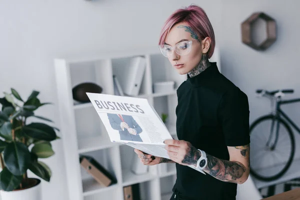 Молода татуйована бізнес-леді в окулярах читає газету на робочому місці — стокове фото
