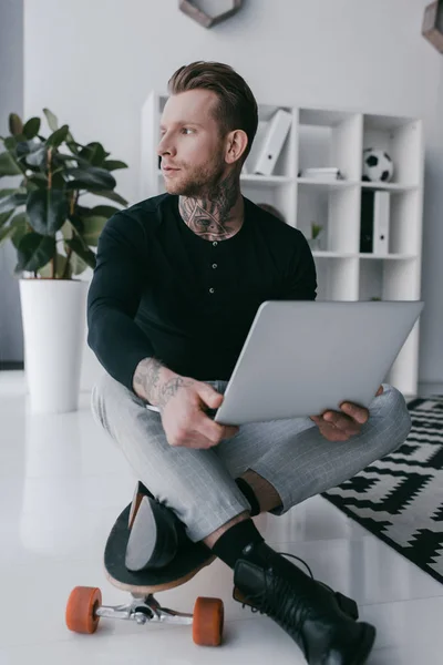 Young businessman sitting on skateboard and using laptop in office — Stock Photo