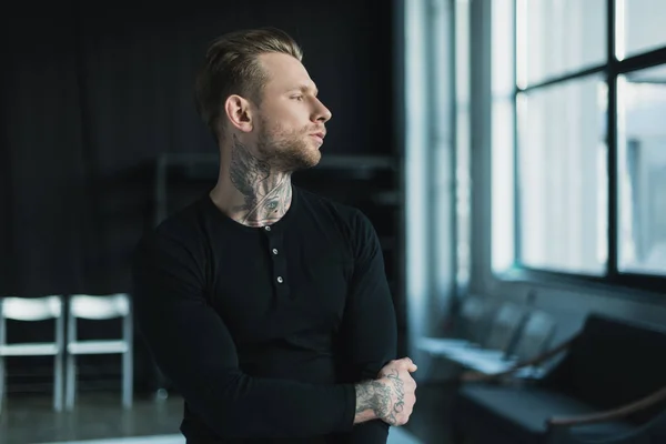 Handsome young man with tattoos standing and looking away indoors — Stock Photo