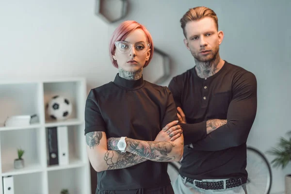Молоді татуйовані бізнесмени стоять з схрещеними руками і дивляться на камеру в офісі — стокове фото