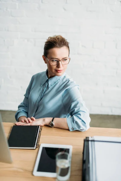 Mujer de negocios adulta elegante sentada en el lugar de trabajo en la oficina y mirando hacia otro lado - foto de stock