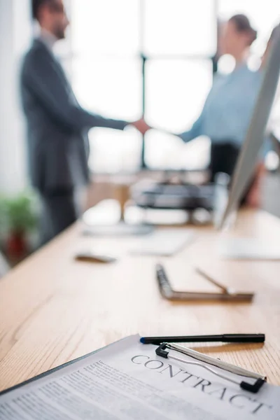 Close-up shot of business contract lying on table and blurred business partners shaking hands on background — Stock Photo