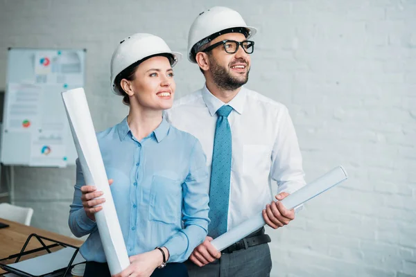 Arquitectos felices de pie en la oficina moderna con planos de edificios y mirando hacia otro lado - foto de stock