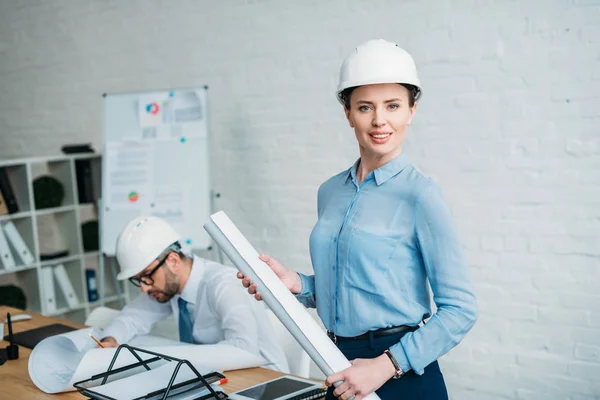 Beautiful female architect standing at modern office with building plan while his colleague working on background — Stock Photo