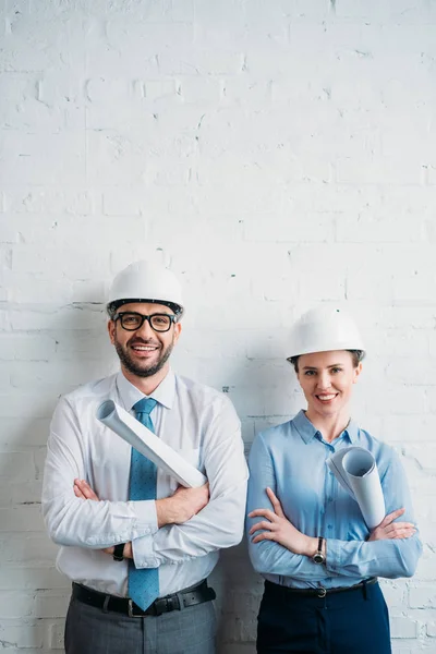 Arquitectos sonrientes en sombreros duros de pie frente a la pared de ladrillo blanco con planos - foto de stock