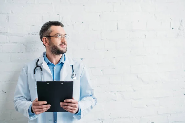 Мужчина врач с планшетом перед стеной из белого кирпича — стоковое фото