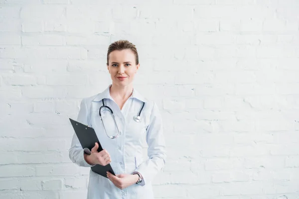 Beautiful female doctor with clipboard in front of white brick wall — Stock Photo