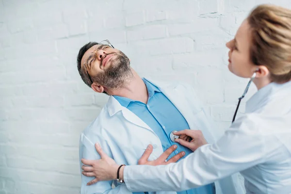 Female doctor listening to heartbeat of colleague who has heart attack with stethoscope — Stock Photo