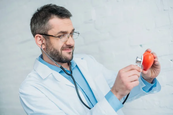Красивий усміхнений лікар слухає серцебиття іграшкового серця зі стетоскопами — стокове фото
