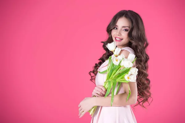 Chica atractiva en vestido rosa con ramo de tulipanes mirando a la cámara aislada en rosa - foto de stock
