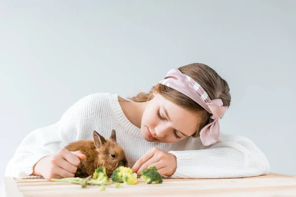 Adorabile ragazza che alimenta simpatico coniglio peloso con broccoli a tavola di legno — Foto stock