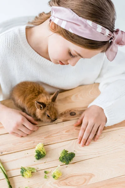 Menina bonita alimentando bonito coelho peludo com brócolis na mesa de madeira — Fotografia de Stock