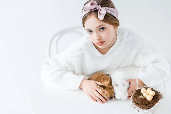 Hermosa chica mirando a la cámara mientras está sentado con conejos peludos y cesta con huevos de Pascua en el nido en blanco - foto de stock