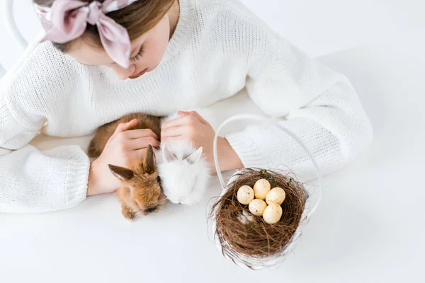 Tiro cortado de menina olhando para coelhos peludos bonitos e cesta com ovos de Páscoa no ninho — Fotografia de Stock