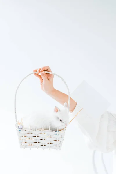 Primer plano vista parcial de niña sosteniendo cesta con lindo conejo peludo y tarjeta en blanco aislado en blanco - foto de stock