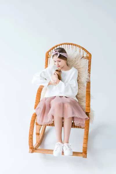 Bella ragazza tenendo peloso coniglio mentre seduto su sedia a dondolo isolato su bianco — Foto stock