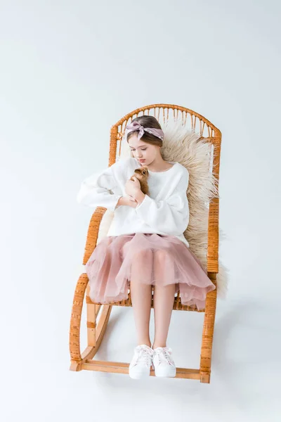 Beautiful girl holding furry rabbit while sitting on rocking chair isolated on white — Stock Photo