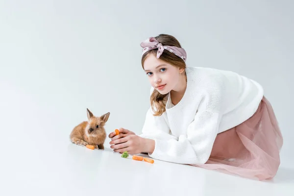 Adorable girl looking at camera while feeding rabbit isolated on white — Stock Photo