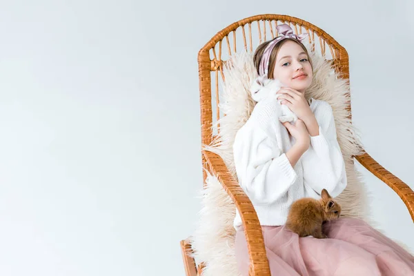 Красива дівчина-підліток посміхається на камеру, сидячи в кріслі-гойдалці з пухнастими кроликами ізольовані на білому — стокове фото