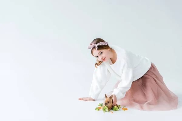 Bella ragazza sorridente alla macchina fotografica mentre alimenta coniglietto isolato su bianco — Foto stock