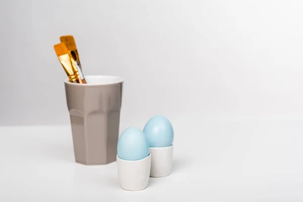 Vista de cerca de huevos de Pascua pintados de azul y pinceles de pintura en taza en gris - foto de stock