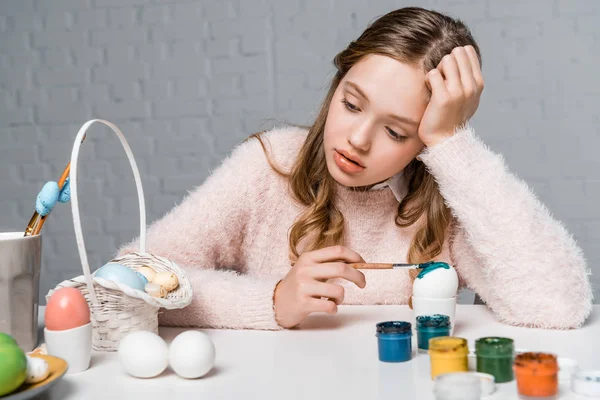Pensive teenage girl painting easter egg and looking at basket — Stock Photo